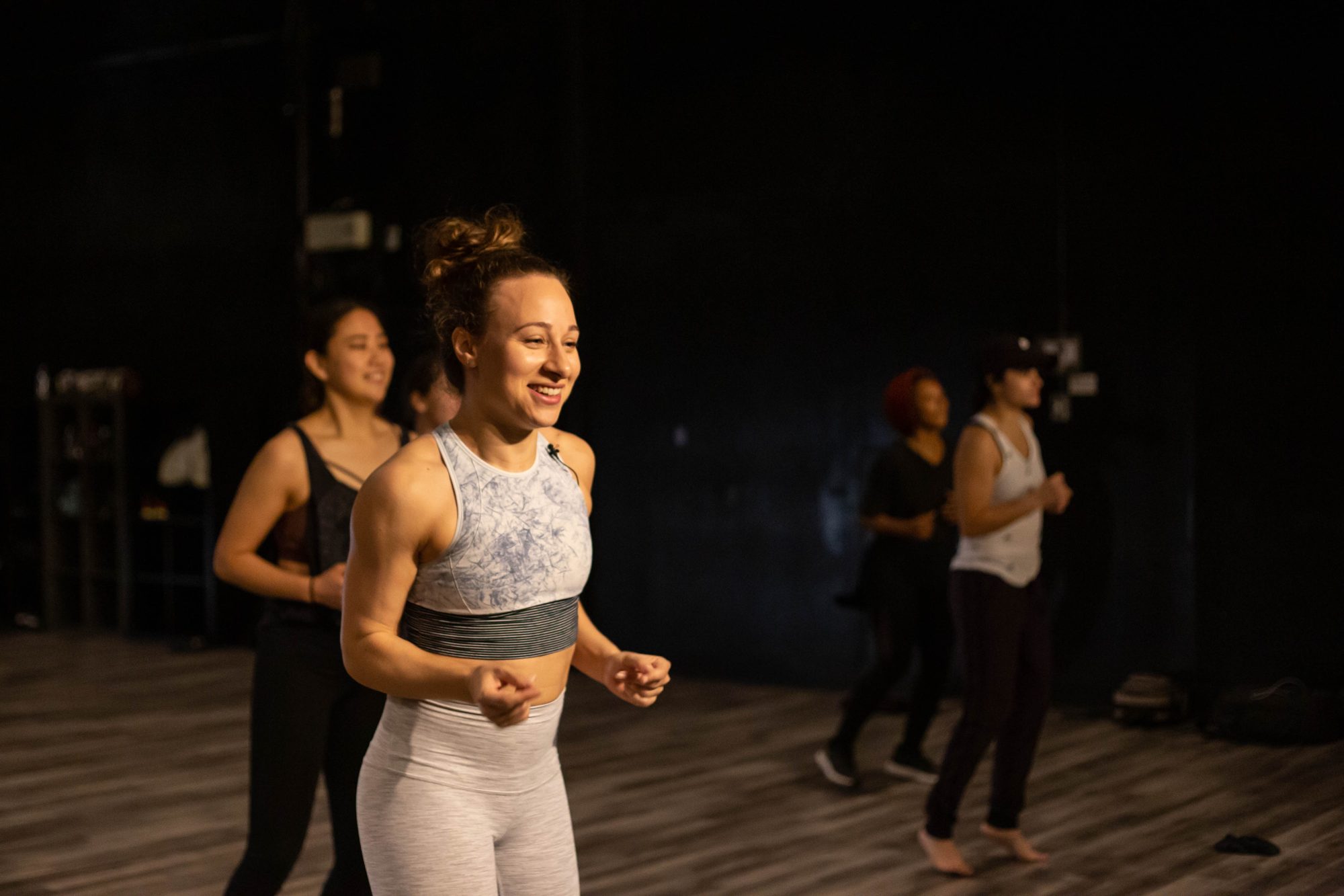Galit smiling at the front of a conditioning class in a dark studio, the class behind her