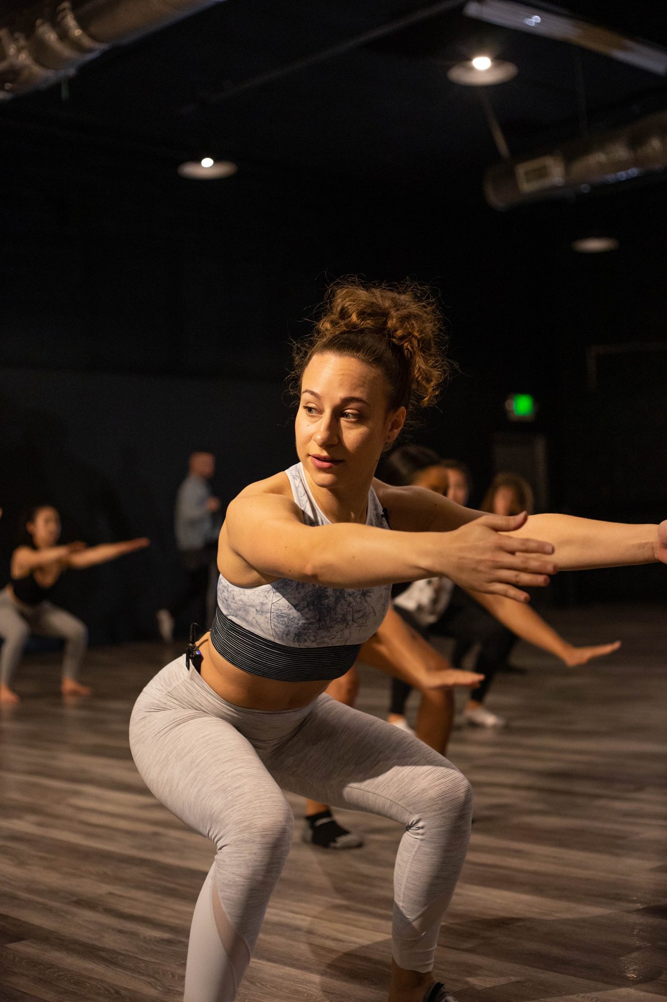 Galit low in a squat with one arm reaching out, the class behind her in the dark studio