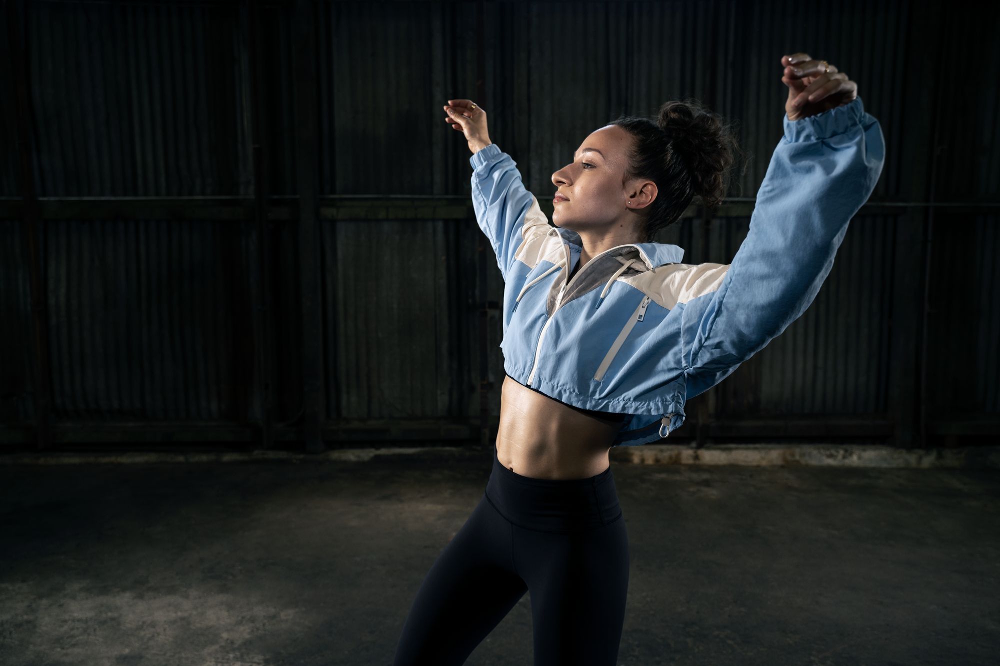 Galit Friedlander mid movement in a dark warehouse, arms thrown wide in a denim jacket