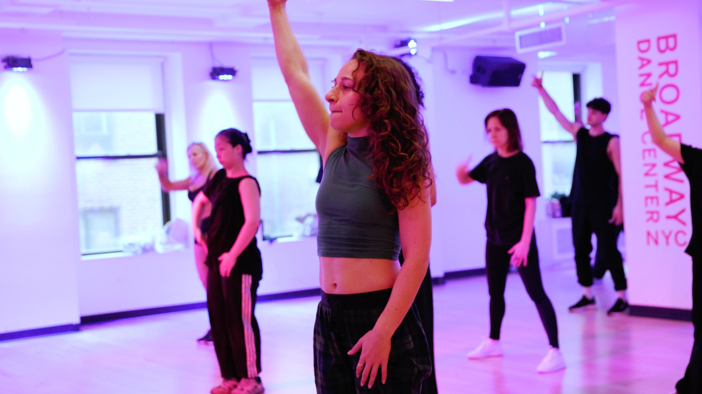 Galit at the front of a class at Broadway Dance Center, one arm up, the room following her