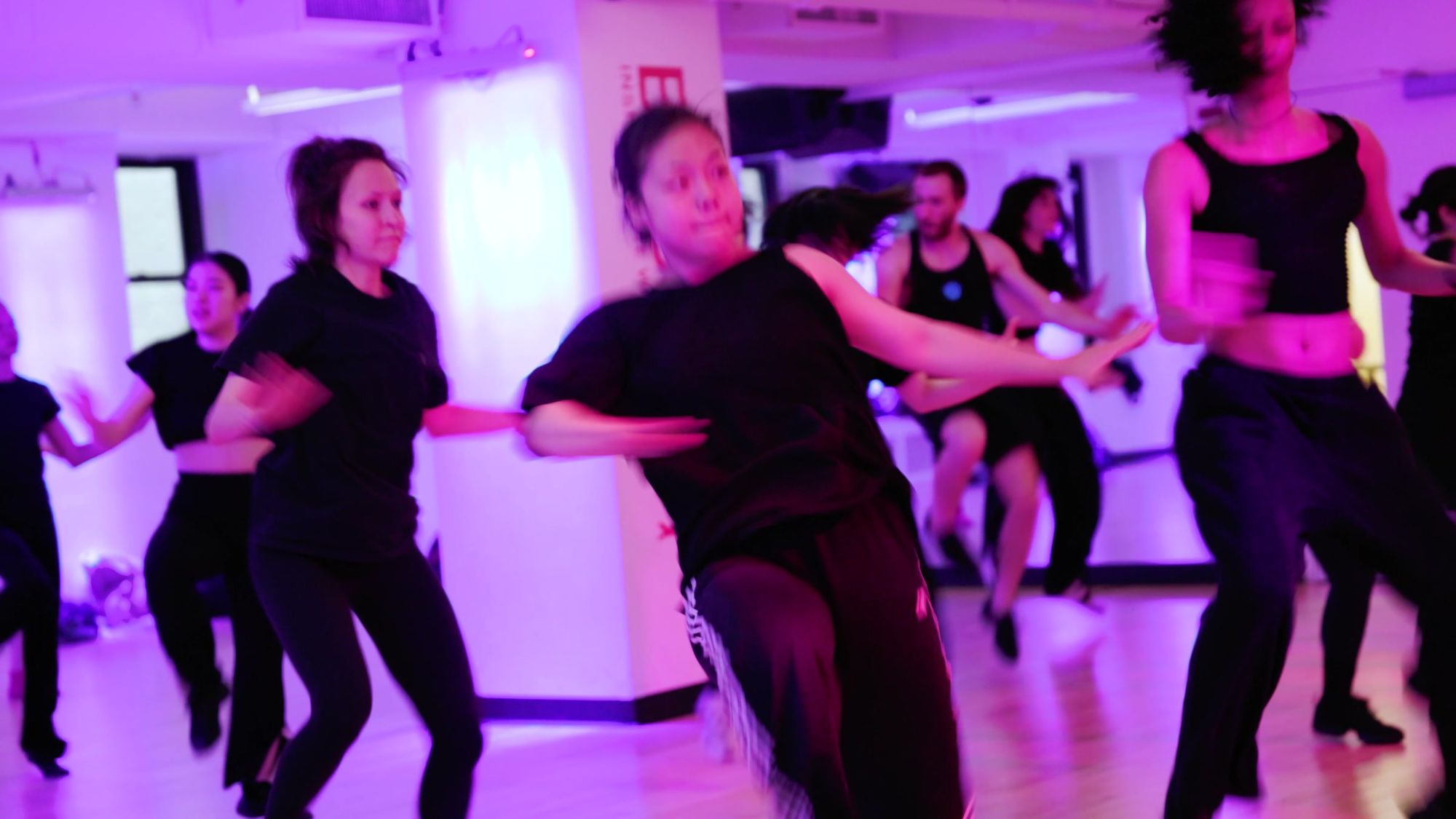 Dancers mid step together under magenta light, arms swinging through the same move